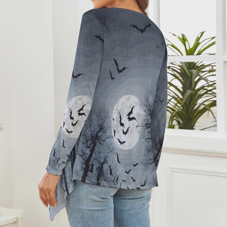 May include: A long-sleeve top featuring a gray and black gradient print of a night sky with two full moons, silhouettes of bare tree branches, and numerous flying bats. The garment has a rounded neckline and a slightly asymmetrical hemline.