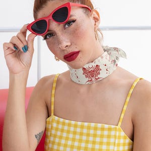 May include: A yellow and white gingham square-neck top paired with a cream-colored neck scarf featuring a red moth and celestial print. The ensemble includes bright red cat-eye sunglasses with dark lenses, creating a bold and vibrant look.