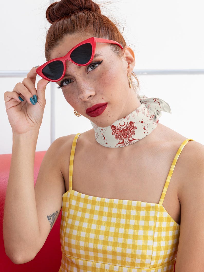 May include: A yellow and white gingham square-neck top paired with a cream-colored neck scarf featuring a red moth and celestial print. The ensemble includes bright red cat-eye sunglasses with dark lenses, creating a bold and vibrant look.