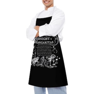 May include: A black apron featuring a white graphic print with the text "MIDNIGHT MARGARITAS" and a poem excerpt. The design includes illustrations of a crescent moon, a frog, a cocktail shaker, a margarita glass, and stars in a white ink style.