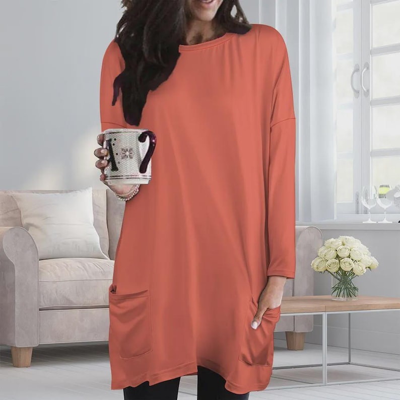 May include: A long-sleeved, solid coral-colored tunic top featuring a round neckline, dropped shoulders, and two large patch pockets on the front. The garment has a relaxed, loose-fitting silhouette that extends to the upper thigh area.