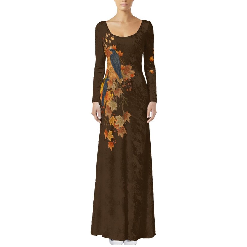 May include: A long-sleeved, floor-length brown dress featuring a scoop neckline. The garment is decorated with a printed design of two dark birds perched among a cluster of vibrant autumn leaves in shades of orange, yellow, and gold.