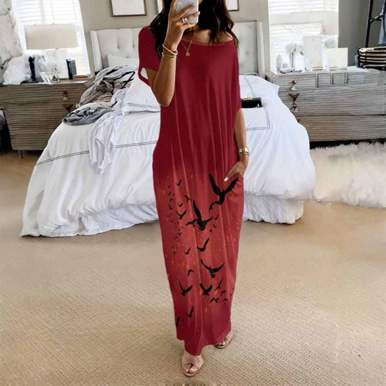 May include: A loose-fitting, short-sleeved maxi dress in a deep red color. The dress features a wide, rounded neckline and a graphic print of multiple black birds in flight scattered across the lower portion of the garment. It includes side pockets.