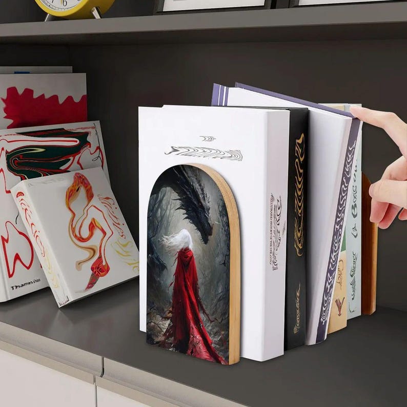 K&ouml;nnte beinhalten: Ein h&ouml;lzerner Buchst&auml;nder mit einem gew&ouml;lbten Einsatz, der eine Illustration einer Figur in einem langen roten Umhang vor einem gro&szlig;en schwarzen Drachen in einem nebligen Wald zeigt. Der Buchst&auml;nder st&uuml;tzt eine Reihe von B&uuml;chern mit wei&szlig;en, schwarzen und beigen Buchr&uuml;cken.