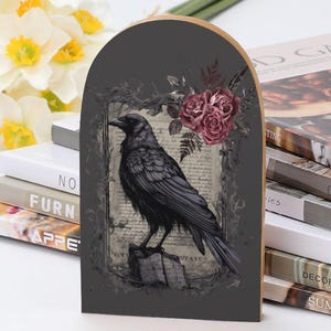 May include: Wooden arched plaque featuring a detailed illustration of a black raven perched on a weathered stump. The raven is set against a dark gray background with vintage floral and script elements. A cluster of burgundy roses adds a touch of color.
