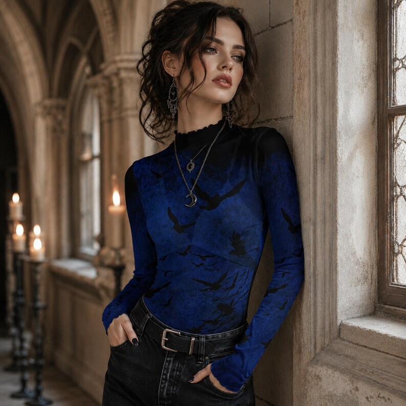 May include: A long-sleeve mock neck top featuring a deep blue and black gradient pattern with silhouettes of flying birds. The garment has a fitted silhouette, a high neckline with a subtle ruffled edge, and long sleeves, paired with black jeans.