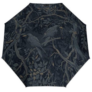 Puede incluir: Un paraguas azul marino oscuro con una ilustración detallada de dos cuervos posados en ramas dentro de un bosque. El diseño incluye hojas y ramas en tonos grises y marrones, creando un tema inspirado en la naturaleza.