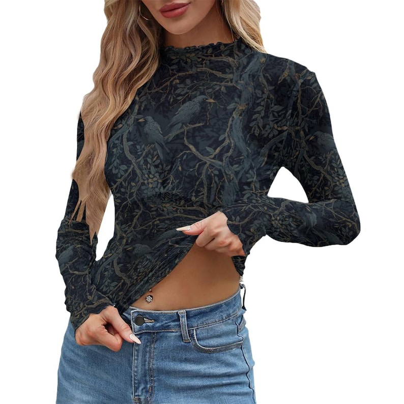 May include: A long-sleeve mock neck top featuring a dark forest print with silhouettes of birds perched on tree branches. The fabric has a subtle texture and a fitted silhouette, paired with blue denim jeans that have a visible metal button closure.