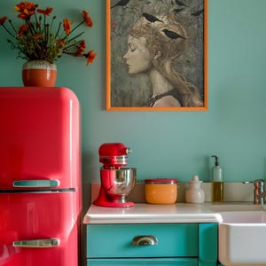 May include: A kitchen scene featuring a bright red retro refrigerator, a red stand mixer, and teal cabinets. A framed print of a person with birds in their hair hangs on the wall above a white sink, next to a vase of orange flowers.