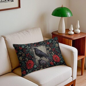 May include: A rectangular throw pillow featuring a dark background with a pattern of red roses and green foliage. In the center, a black crow is perched on a stack of books within a rectangular frame that contains faint, illegible text.
