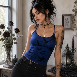 May include: A blue tank top featuring a black bird silhouette print, paired with black denim jeans. The tank top has thin spaghetti straps and a scoop neckline. The garment is worn with a layered necklace set and a black lace choker accessory.