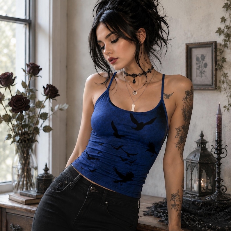 May include: A blue tank top featuring a black bird silhouette print, paired with black denim jeans. The tank top has thin spaghetti straps and a scoop neckline. The garment is worn with a layered necklace set and a black lace choker accessory.