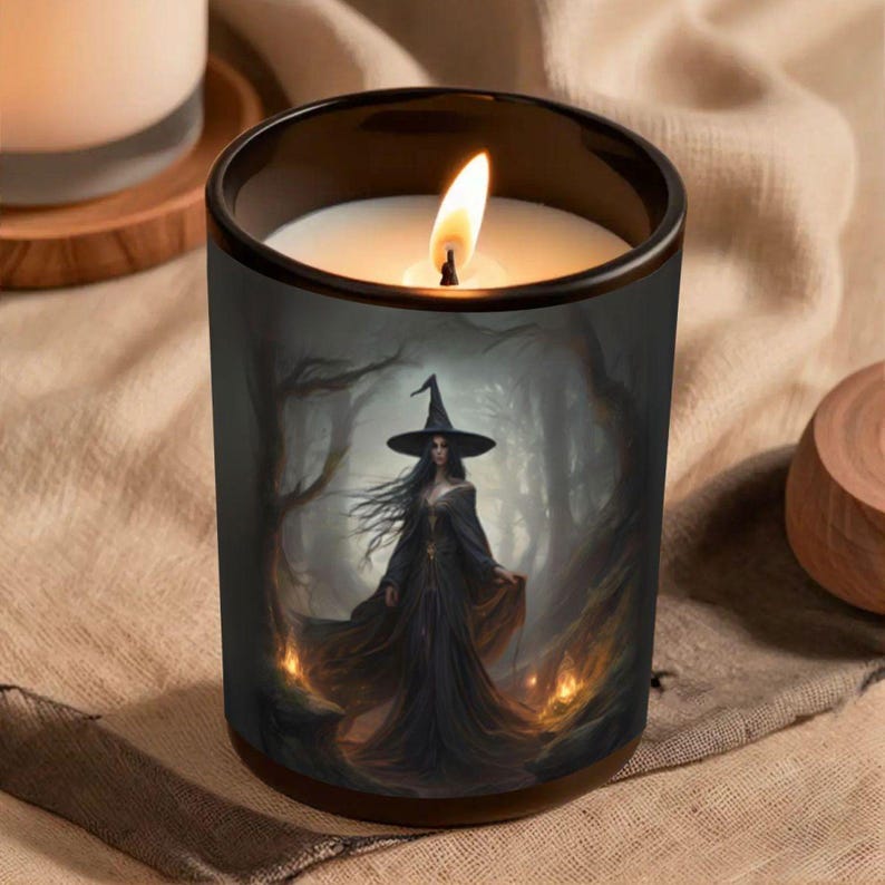 May include: A burning candle in a black glass jar. The jar is decorated with a fantasy image of a witch in a black dress and hat, set in a dark forest with flames. The candle is white, and the flame is bright.