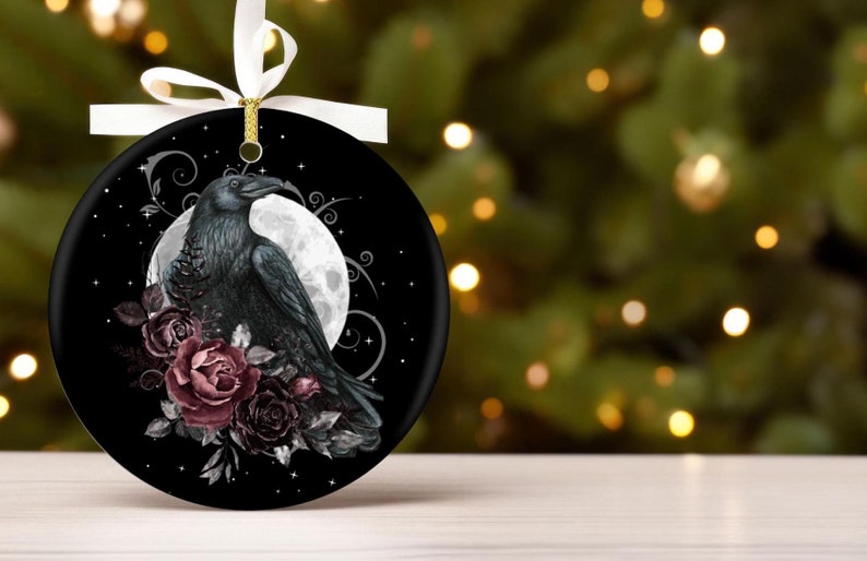 May include: A round black ceramic ornament featuring a detailed illustration of a black raven perched before a large white full moon. The design includes dark red roses and swirling silver filigree accents. A white ribbon is tied at the top.