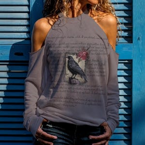 May include: A grey long-sleeve cold-shoulder top featuring a graphic of a black raven perched on a book with pink roses. The fabric is printed with the text of Edgar Allan Poe's The Raven, including the lines Once upon a midnight dreary.