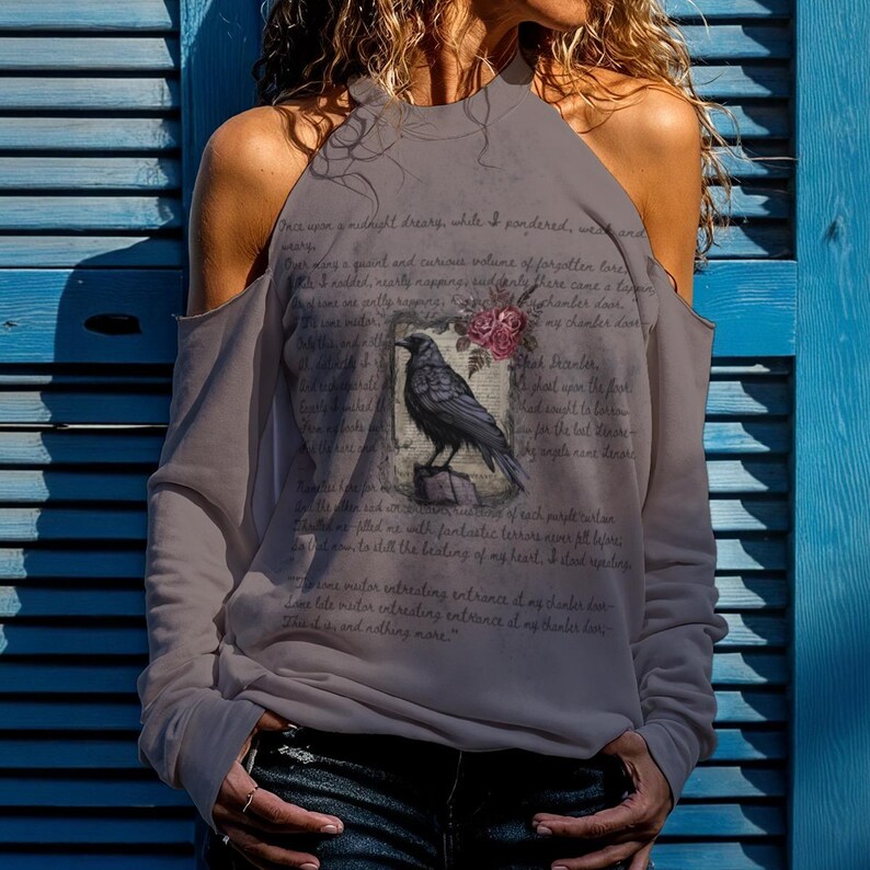 May include: A grey long-sleeve cold-shoulder top featuring a graphic of a black raven perched on a book with pink roses. The fabric is printed with the text of Edgar Allan Poe's The Raven, including the lines Once upon a midnight dreary.