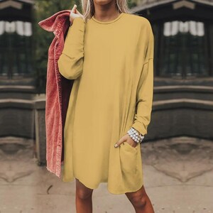 May include: A mustard yellow long-sleeve tunic dress with a round neckline and a front pocket. The dress features a loose, relaxed fit and is paired with a textured, dark pink corduroy jacket draped over one shoulder, creating a layered look.