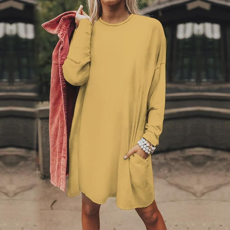 May include: A mustard yellow long-sleeve tunic dress with a round neckline and a front pocket. The dress features a loose, relaxed fit and is paired with a textured, dark pink corduroy jacket draped over one shoulder, creating a layered look.