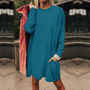 May include: A solid teal long-sleeve dress featuring a round neckline, a relaxed fit, and a single visible patch pocket on the front. A separate rust-colored corduroy jacket is draped over one shoulder, adding a contrasting texture and color.