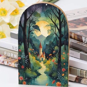 May include: Wooden arched wall art featuring a watercolor painting of a fairytale castle in a forest. The artwork depicts a full moon, a river, and colorful flowers. The castle has red-roofed towers.