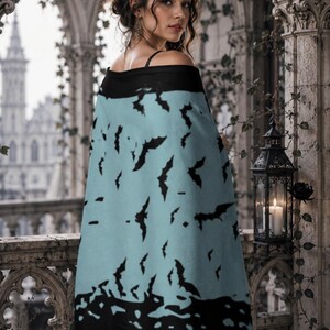 May include: A light blue rectangular shawl featuring a repeating pattern of black bat silhouettes. The shawl has a solid black border along the top and bottom edges, providing a distinct contrast to the central light blue section with bats.