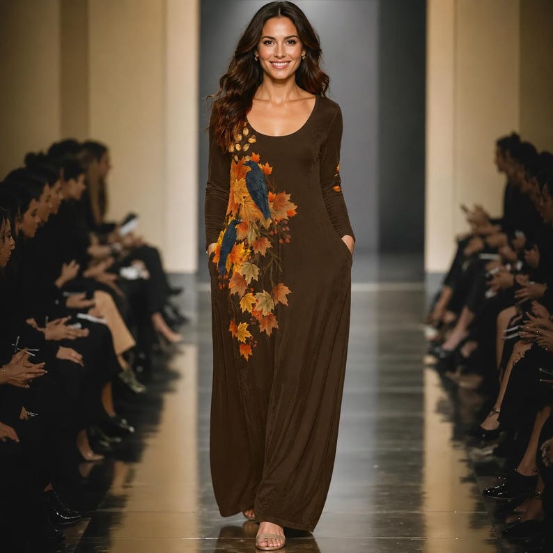 May include: A long-sleeved, floor-length brown dress featuring a scoop neckline and side pockets. The front of the dress is decorated with a printed design of two dark birds perched on a branch surrounded by vibrant orange, yellow, and red maple leaves.