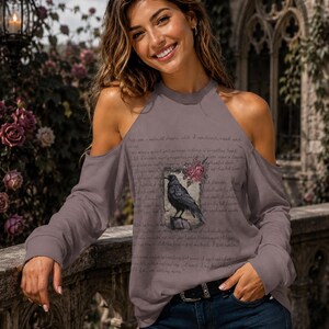 May include: A mauve cold-shoulder long-sleeve top featuring a graphic print of a black raven perched on a wooden post, surrounded by pink roses and overlaid with cursive script text that reads Once upon a midnight dreary while I pondered weak and weary.