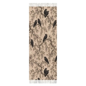 Op de afbeelding: Een rechthoekige sjaal met een beige achtergrond en een herhalend patroon van zwarte vogels op donkerbruine boomtakken. Het ontwerp bevat kleine halve maantjes verspreid tussen het gebladerte en is afgewerkt met franjes aan de uiteinden.