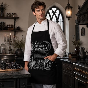 May include: A black apron featuring a white graphic print with the text Midnight Margaritas, a crescent moon, stars, a cocktail shaker, a margarita glass, and a frog illustration, worn over a white long-sleeved chef coat in a kitchen.
