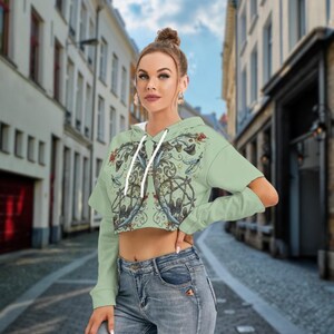 May include: A light green cropped hoodie featuring long sleeves with shoulder cutouts, a drawstring hood, and a detailed graphic print on the front depicting mushrooms, swirling vines, and small stars in earthy tones, paired with blue denim jeans.