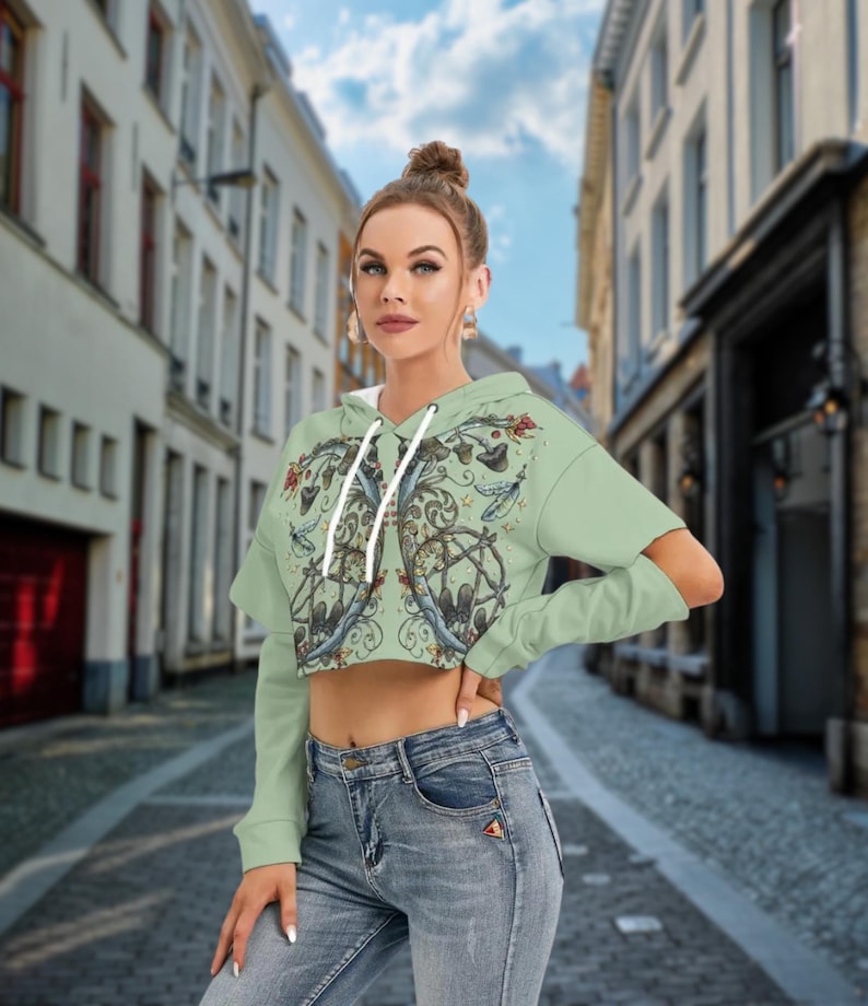 May include: A light green cropped hoodie featuring long sleeves with shoulder cutouts, a drawstring hood, and a detailed graphic print on the front depicting mushrooms, swirling vines, and small stars in earthy tones, paired with blue denim jeans.