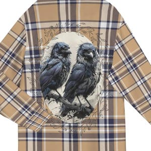 May include: A tan and blue plaid flannel shirt with a circular design featuring two black ravens perched on a branch. The design is surrounded by a wreath of leaves.