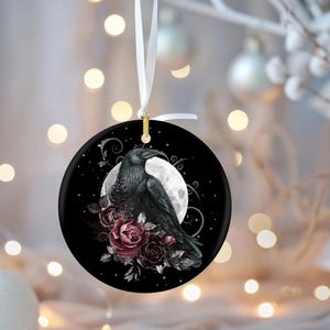 May include: A round ceramic ornament featuring a black raven perched in front of a full moon, surrounded by dark red and purple roses. The design is set against a black background with swirling silver accents and hangs from a white ribbon.
