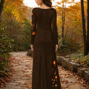 May include: A floor-length dark brown long-sleeve dress featuring a subtle velvet-like texture. The garment is adorned with embroidered orange and yellow autumn leaf motifs scattered along the right sleeve and the lower left side of the skirt.