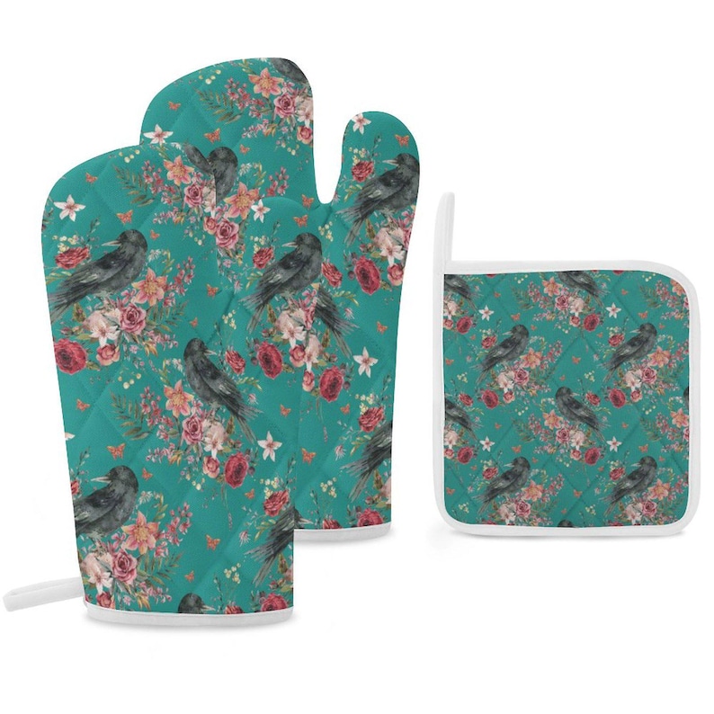 May include: A quilted oven mitt and a square pot holder featuring a teal background with a repeating pattern of black crows, deep red roses, and light pink flowers. Both items have white trim and a small loop for hanging on the side of the pieces.