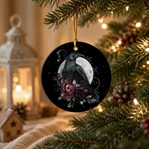 May include: A round ceramic ornament featuring a black raven perched in front of a full moon, surrounded by dark red roses and swirling decorative lines. The ornament is black with a glossy finish and hangs from a thin gold cord.