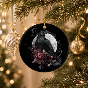 May include: A round black ceramic ornament featuring a detailed illustration of a black raven perched in front of a large white full moon. The raven is surrounded by deep red and dark purple roses with swirling vine accents and small stars.