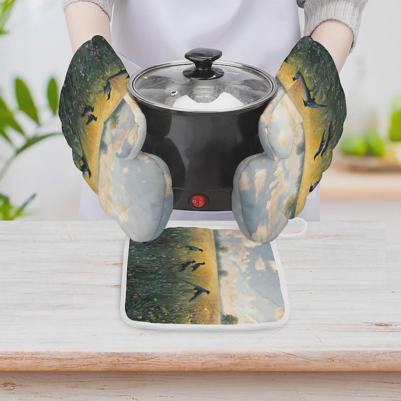 May include: A pair of quilted oven mitts and a matching square pot holder featuring a printed landscape scene of birds flying over a golden field under a cloudy sky. The mitts are shown holding a black cooking pot with a clear glass lid.