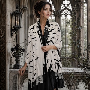 May include: A white shawl featuring a repeating black bat pattern and a solid black border. The garment has long, thin fringe detailing along the bottom edge and is draped over a solid black dress, creating a high-contrast monochromatic look.