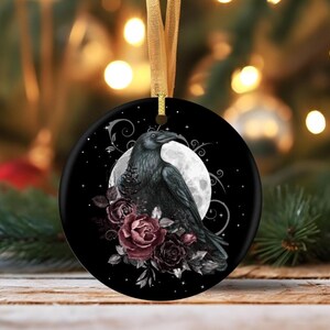 May include: A round black ceramic ornament featuring a detailed illustration of a black raven perched before a full moon, accented by deep red and dark purple roses, hanging from a gold ribbon against a blurred festive holiday background.