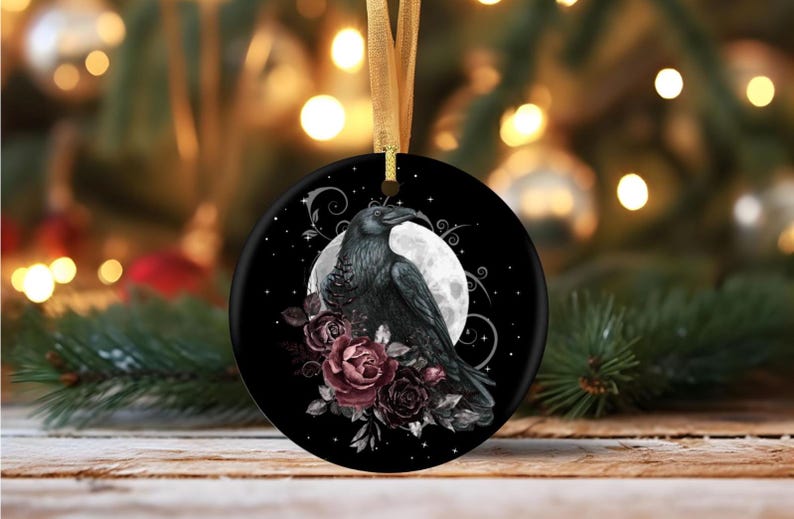 May include: A round black ceramic ornament featuring a detailed illustration of a black raven perched before a full moon, accented by deep red and dark purple roses, hanging from a gold ribbon against a blurred festive holiday background.