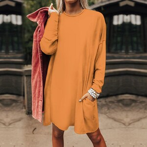 May include: A solid orange long-sleeve dress with a round neckline and a single visible pocket on the front. A textured maroon corduroy jacket is draped over one shoulder, adding a contrasting layer to the plain orange garment.