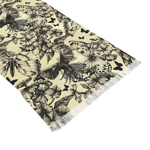 Peut inclure: Un chemin de table rectangulaire au fond cr&egrave;me, orn&eacute; d'une illustration &agrave; l'encre noire repr&eacute;sentant des corbeaux en vol, divers papillons et des grappes de fleurs. Les bords du chemin de table sont finis avec une frange blanche.