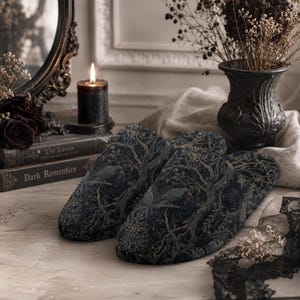 May include: A pair of slip-on slippers featuring a dark, intricate pattern of ravens perched on gnarled, leafless tree branches. The design uses muted tones of charcoal, deep blue, and subtle gold highlights to depict a forest scene on the fabric.