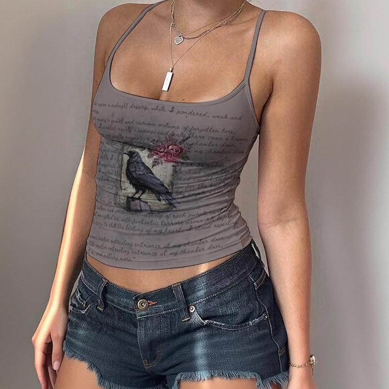 May include: A grey sleeveless vest top featuring a graphic print of a black raven perched on a branch with red roses. The fabric is covered in cursive script text. The top has thin spaghetti straps and a scoop neckline, paired with denim shorts.
