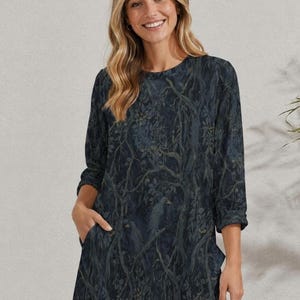 May include: A dark navy blue dress featuring a long-sleeve design with a round neckline and a repeating pattern of forest branches and leaves in muted tones. The garment includes functional side pockets and a straight hemline reaching mid-thigh.