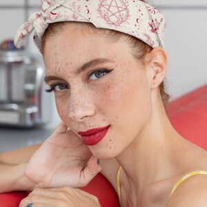 May include: A cream-colored hair bandana featuring a repeating pattern of dark red botanical illustrations, crescent moons, stars, and a central pentagram symbol. The fabric is tied in a knot at the top, creating a wide headband style for hair.