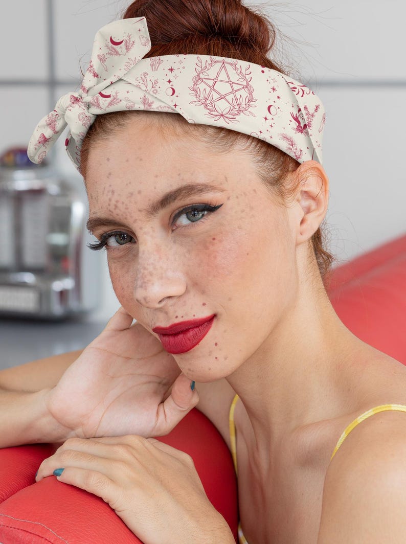 May include: A cream-colored hair bandana featuring a repeating pattern of dark red botanical illustrations, crescent moons, stars, and a central pentagram symbol. The fabric is tied in a knot at the top, creating a wide headband style for hair.