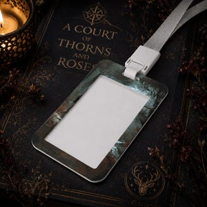 Puede incluir: Un portatarjetas de identificaci&oacute;n rectangular con un estampado de bosque oscuro y atmosf&eacute;rico en tonos azules y marrones apagados. El soporte incluye un conector de pl&aacute;stico blanco unido a un cord&oacute;n de tela gris claro para tarjetas.