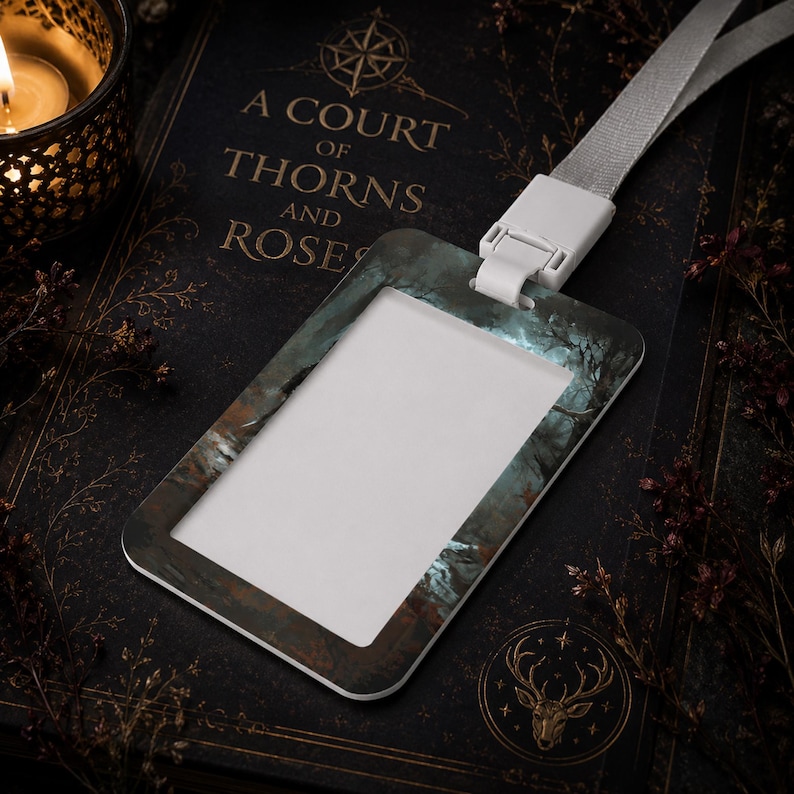 Puede incluir: Un portatarjetas de identificaci&oacute;n rectangular con un estampado de bosque oscuro y atmosf&eacute;rico en tonos azules y marrones apagados. El soporte incluye un conector de pl&aacute;stico blanco unido a un cord&oacute;n de tela gris claro para tarjetas.